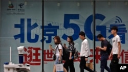 FILE - People walk toward a police robot mounted with surveillance cameras patrol past a 5G network advertisement at a shopping district in Beijing, May 15, 2019. 