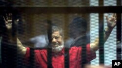 Mantan Presiden Mesir Mohammed Morsi, mengenakan jumpsuit merah (baju bagi napi yang dijatuhi hukuman mati), mengangkat tangannya di dalam "kandang terdakwa" saat menghadiri sidang di akademi kepolisian nasional, di pinggiran timur Kairo, Mesir, 18 Juni 2016. (Foto: dok).