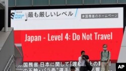 People wearing masks to help protect against the spread of the coronavirus walk in front of a screen showing the news on U.S. warning against visits to Japan, May 25, 2021, in Tokyo. 