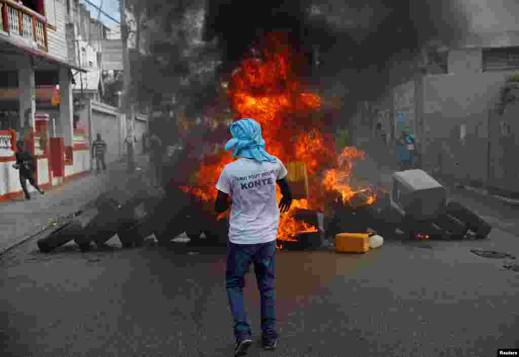 Yon manifestan mete yon mayo ki di "lavi tout moun KONTE" pandan li kanpe devan yon barikad kap boule.