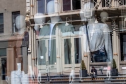 A woman sitting on a stoop reading a book in the sun is seen reflected by a closed clothing store's window on West Broadway on May 7, 2020, in the SoHo neighborhood of the Manhattan borough in New York.