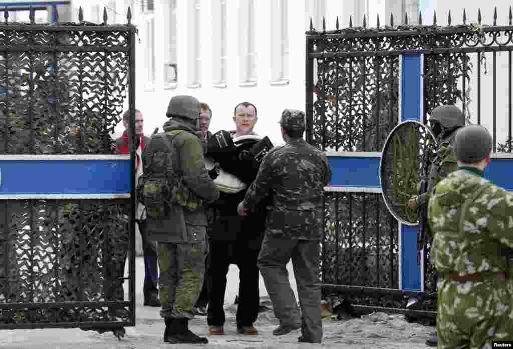 A Ukrainian naval officer carries his belongings as he walks out of the territory of the naval headquarters, with armed men, believed to be Russian servicemen, seen nearby, in Sevastopol, March 19, 2014.