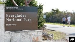 Visitors walk past a sign for Everglades National Park as they enter from overflow parking, Jan. 2, 2019, in Everglades National Park, Fla. Human feces, overflowing garbage, illegal off-roading and other damaging behavior were beginning to to overwhelm some of the national parks, as a partial government shutdown drags on.