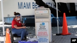 A poll worker wears personal protective equipment as she monitors a ballot drop box for mail-in ballots outside of a polling station during early voting, Aug. 7, 2020, in Miami Beach, Florida. 