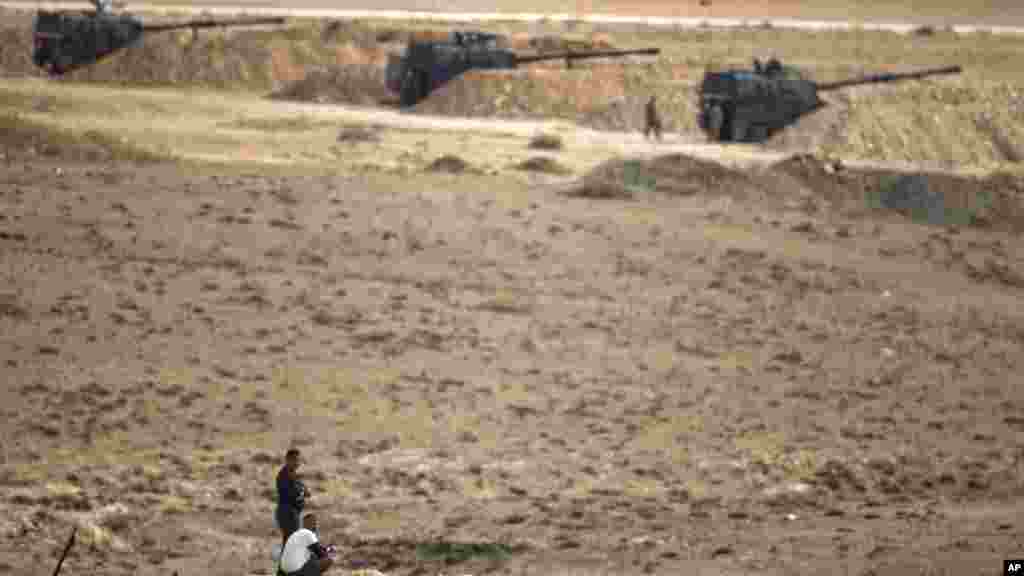 Men watch fighting across the border in Kobani from a hilltop on the outskirts of Suruc, Turkey, backdropped by Turkish army artillery positions near the Turkey-Syria border, Oct. 26, 2014.