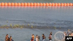 Sejumlah migran berjalan di antara kawat berduri dan pelampung di sepanjang perbatasan antara AS-Meksiko di sungai Rio Grande di Eagle Pass, Texas, pada 16 Juli 2023. (Foto: AFP/Suzanne Cordeiro)