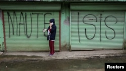 FILE - A woman is seen using a mobile phone while standing in front of a shuttered storefront with "MAUTE-ISIS" graffiti, in Marawi city, southern Philippines, Oct. 20, 2017. Maute, an Islamic State affiliate, was among groups the U.S. added to its terror list Tuesday.